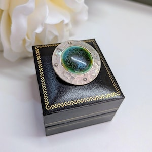 May include: A circular brooch featuring a central, glossy, dome-shaped cabochon with a gradient of deep green and blue hues. The cabochon is set within a hammered, textured silver-toned metal frame accented with four small, evenly spaced rivets.