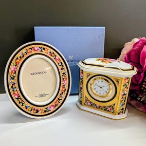 Puede incluir: Un marco de fotos ovalado y un reloj de cuarzo rectangular de Wedgwood, ambos con un borde negro y un patrón floral y de frutas colorido. El marco muestra texto para un tamaño de foto de 65mm por 75mm. Una caja azul se sitúa detrás.
