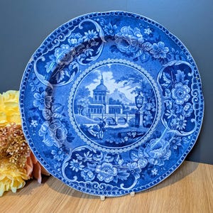 May include: A round ceramic plate featuring a blue and white transferware pattern. The centre depicts a landscape with a building, a bridge, and figures. The border is decorated with floral motifs and scrollwork, finished with a scalloped edge.