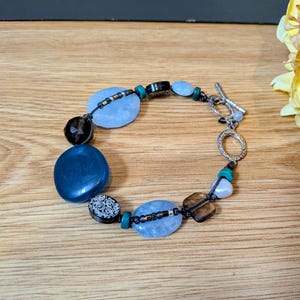 May include: A bracelet featuring a mix of blue, black, and teal beads, including large smooth blue stones, faceted dark brown beads, and a black bead with white scroll patterns. It is finished with a silver-toned metal toggle clasp closure.