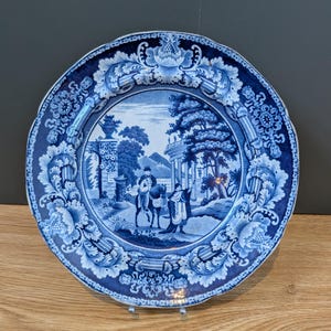 Könnte beinhalten: Ein runder Keramikteller mit einem blau-weißen Transferdruckmuster. Die Mitte zeigt eine Landschaft mit einer Person auf einem Pferd und einer weiteren stehenden Figur bei antiken Ruinen. Der Rand ist mit floralen Ornamenten verziert.
