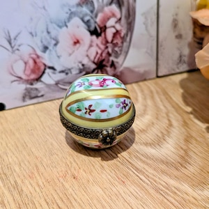 May include: A small, spherical porcelain trinket box with a pale yellow base and white panels. The surface features hand-painted pink and purple floral motifs with green leaves and gold-toned horizontal bands. It has an ornate metal hinge.