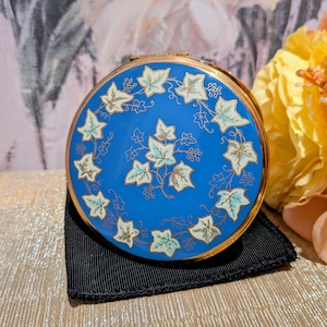 May include: A round, gold-toned metal compact mirror featuring a vibrant blue enamel lid. The lid is decorated with a central cluster of light green ivy leaves and a circular border of matching ivy vines and small berries in a delicate gold outline.