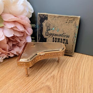 Puede incluir: Una caja de música en miniatura con forma de piano de cola en metal dorado, con un intrincado patrón grabado en la tapa y los laterales. Se coloca frente a una caja de cartón cuadrada etiquetada como Pygmation SONATA con notas musicales.