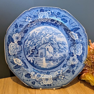 May include: A round ceramic plate with a scalloped edge featuring a blue and white transferware pattern. The center depicts a stone cottage by a water wheel and trees. The wide border is decorated with detailed floral motifs and roses.