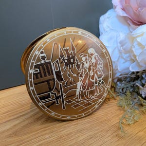 May include: A round, metallic gold-toned tin container featuring a white etched illustration on the lid. The design depicts a horse-drawn carriage, a castle in the background, and two figures standing together. The lid has a decorative border.