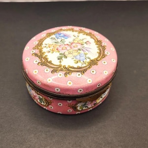May include: A round pink metal trinket box featuring a white center medallion with a colorful floral bouquet surrounded by a gold-toned ornate frame. The exterior surface is decorated with a repeating pattern of small white and gold daisy-like flowers.