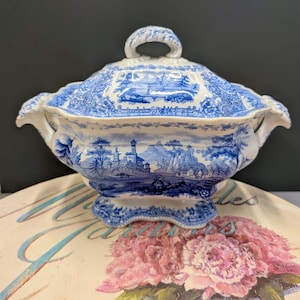 May include: A white ceramic tureen with a blue transferware pattern featuring a landscape with castles, trees, and mountains. The lidded serving dish has two side handles, a scalloped base, and a decorative handle on the lid.