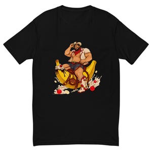 May include: A black t-shirt featuring a graphic illustration of a bearded person wearing a cowboy hat, red bandana, and white vest, riding a large yellow banana like a horse. The banana is topped with chocolate sauce, whipped cream, and cherries.
