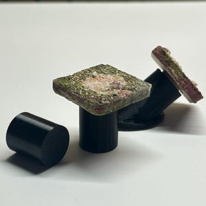 May include: Three black cylindrical stands, two of which are topped with square, textured, moss-like green and pink decorative pieces. The third stand is plain black and cylindrical, positioned separately on the white surface.