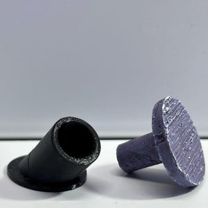 May include: Two small, cylindrical components with flat, circular bases. One component is matte black with a hollow center, while the other is a textured, metallic purple color with a solid, flat top surface. Both items feature visible 3D printed layers.