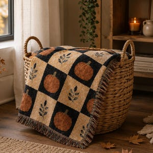 May include: A woven throw blanket featuring a checkered pattern of black and tan squares. The tan squares display orange pumpkins and dark leaf sprigs. The blanket has a fringed edge and is draped over a large, round, woven wicker basket.
