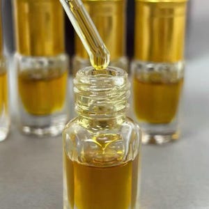 Puede incluir: Un frasco de vidrio transparente con tapa dorada que contiene un líquido de color ámbar. Un gotero de vidrio está colocado sobre el frasco abierto, con una gota de aceite dorado suspendida en la punta. Varios frascos idénticos están dispuestos al fondo.