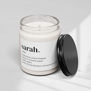 Puede incluir: Una vela en frasco de vidrio transparente con cera blanca y tapa de metal negro. La etiqueta blanca presenta el texto: sarah. noun professional online shopper always 5 minutes away coffee addict queen of bad decisions and great stories.