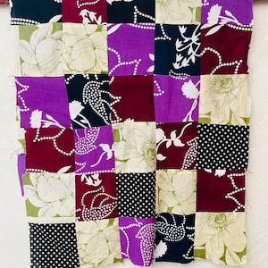 May include: A patchwork quilt featuring a grid of square fabric panels in various patterns and colors, including deep purple, maroon, black with white polka dots, and cream with large green floral prints, all stitched together in a uniform layout.