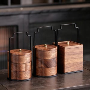 May include: Three tiered wooden food containers with dark brown wood grain patterns. Each container features a black metal handle frame and a small brass-colored knob on the lid. Two containers are cylindrical, and one is square in shape.