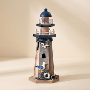 May include: A wooden lighthouse figurine featuring a weathered brown finish, a dark blue roof, and white railings. The structure is decorated with a small white seagull, a larger seagull with blue wings, a white life preserver, and black rope accents.