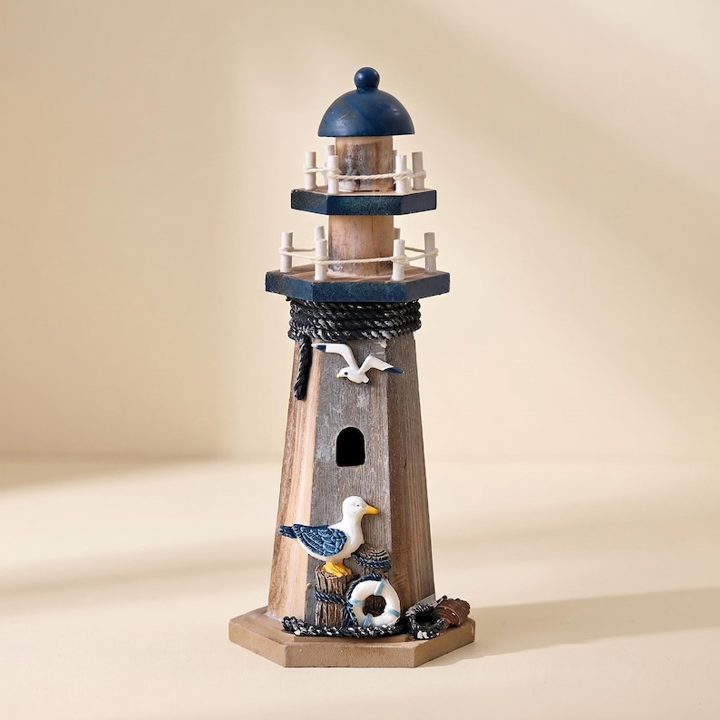 May include: A wooden lighthouse figurine featuring a weathered brown finish, a dark blue roof, and white railings. The structure is decorated with a small white seagull, a larger seagull with blue wings, a white life preserver, and black rope accents.