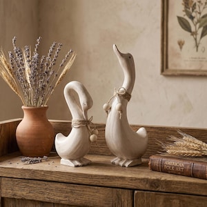 May include: Two off-white duck figurines with distressed paint finishes, each featuring a jute twine neck tie with wooden beads. The ducks are positioned next to a terracotta vase filled with dried lavender and wheat stalks, and a book titled American Folk Art.