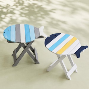 May include: Two folding stools shaped like fish. The left stool has a grey base and a top with blue, white, and grey stripes. The right stool has a white base and a top with dark blue, light blue, white, and yellow stripes. Both have a cutout eye.