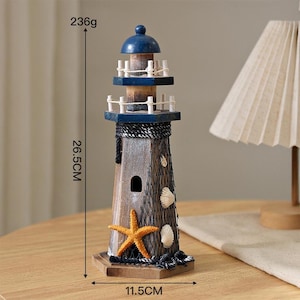 May include: A wooden lighthouse figurine featuring a blue roof, a dark brown body with a black fishing net overlay, a yellow starfish, and white seashells. It measures 26.5 cm in height, 11.5 cm in width, and weighs 236 grams as indicated by text.