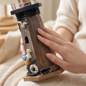 May include: A wooden lighthouse figurine featuring a dark blue top, a black rope accent, and a small arched window. The structure is decorated with a white and blue seagull, a white life preserver, and a small shell at the base on a square stand.