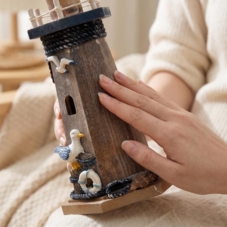 May include: A wooden lighthouse figurine featuring a dark blue top, a black rope accent, and a small arched window. The structure is decorated with a white and blue seagull, a white life preserver, and a small shell at the base on a square stand.