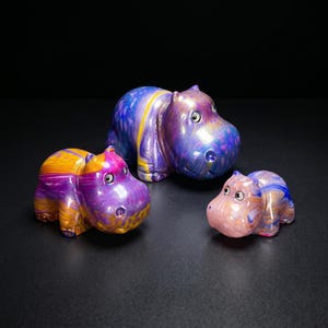 May include: Three hippopotamus figurines crafted from smooth, polished material. The largest figure features a swirling blend of purple, blue, and yellow. The two smaller figures display similar marbled patterns in shades of purple, orange, and light pink.