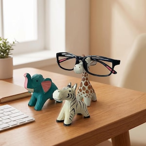 May include: Three hand-carved wooden animal figurines including a teal elephant with pink ears, a white and black striped zebra, and a giraffe with brown spots. The giraffe figurine features a notched snout designed to hold a pair of black eyeglasses.