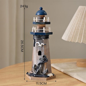 May include: A wooden lighthouse figurine featuring a blue top, two railed balconies, a white seagull cutout, and a black anchor wrapped in rope. It measures 26.5 cm in height, 11.5 cm in width, and weighs 243 grams as indicated by the visible text.