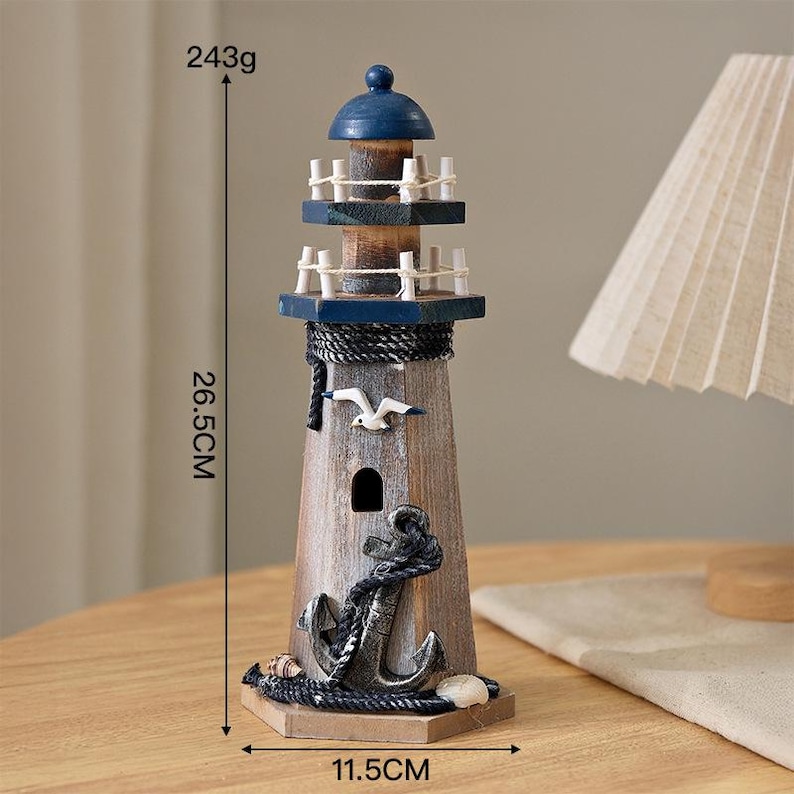 May include: A wooden lighthouse figurine featuring a blue top, two railed balconies, a white seagull cutout, and a black anchor wrapped in rope. It measures 26.5 cm in height, 11.5 cm in width, and weighs 243 grams as indicated by the visible text.