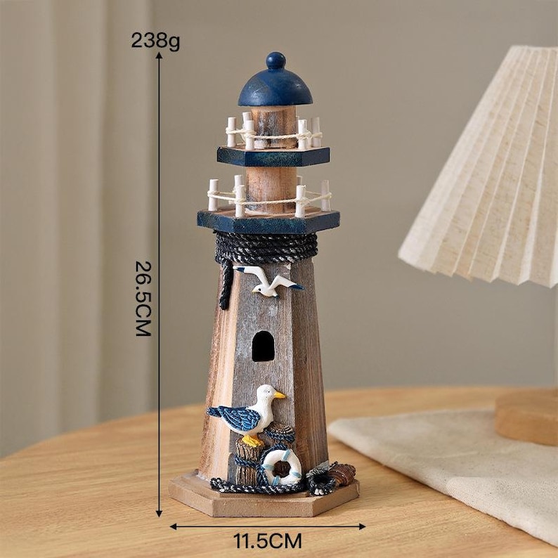 May include: A wooden lighthouse figurine featuring a blue dome top, white railings, and a dark rope accent. The structure is decorated with a seagull and a life ring. It measures 26.5 cm in height, 11.5 cm in width, and weighs 238 grams.
