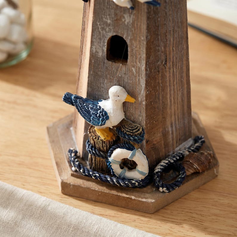 May include: A wooden lighthouse figurine featuring a weathered brown finish, a small arched window, and a decorative base. The base is adorned with a white and blue seagull perched on a post, a white life ring, a coiled navy rope, and a seashell.