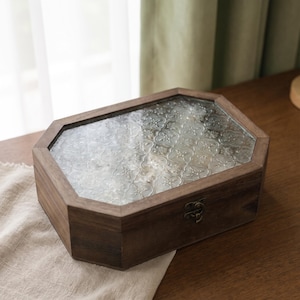 Könnte beinhalten: Eine rechteckige Aufbewahrungsbox aus Holz mit achteckiger Form und Klappdeckel. Der Deckel verfügt über eine klare Glasscheibe mit einem sich wiederholenden, geprägten Blumenmuster. Ein kleiner, dunkler Metallverschluss ist vorne mittig angebracht.