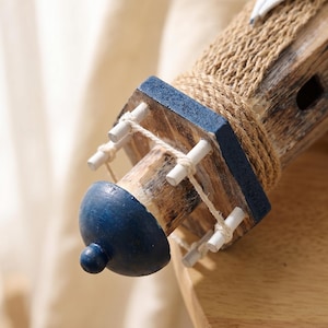 May include: A decorative lighthouse model featuring a weathered brown wood finish, a dark blue painted top, and a wrap of natural tan jute rope. The structure includes a small balcony with white wooden railings connected by thin white string.