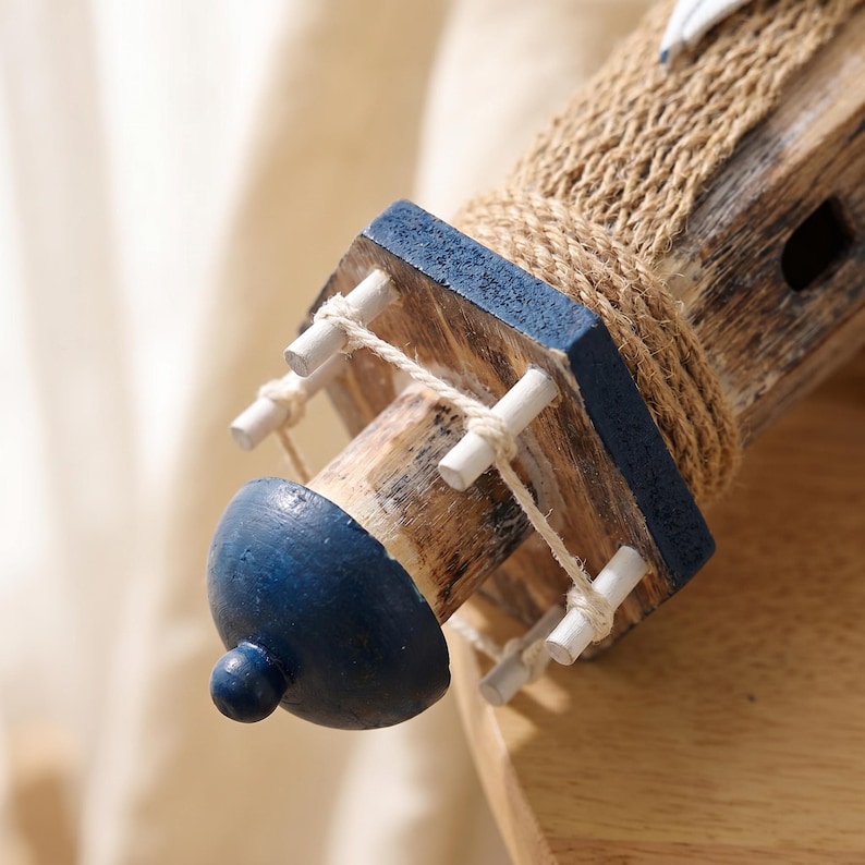 May include: A decorative lighthouse model featuring a weathered brown wood finish, a dark blue painted top, and a wrap of natural tan jute rope. The structure includes a small balcony with white wooden railings connected by thin white string.