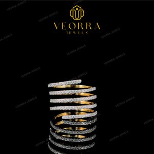 May include: A gold spiral ring featuring multiple bands encrusted with small, clear, sparkling stones. The ring is displayed against a black background with the gold text VEORRA JEWELS and a logo positioned above the jewellery piece.