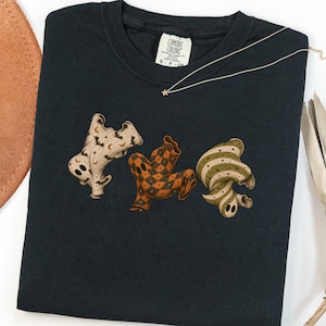 May include: A black short-sleeved t-shirt featuring a graphic of three ghosts. The left ghost has a cream background with black bats and moons. The center ghost has an orange and black diamond pattern. The right ghost has a green and cream striped pattern.