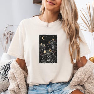 Könnte beinhalten: Ein cremefarbenes Kurzarm-T-Shirt mit einem rechteckigen Grafikdruck von drei als Vampiren verkleideten Fröschen. Die Frösche haben große gelbe Augen und tragen schwarze Umhänge mit hohem Kragen vor einem dunklen Nachthimmel mit Mondsichel.