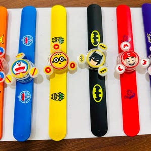 Pode incluir: Seis pulseiras de bater coloridas com vários personagens de desenhos animados, incluindo Pikachu, Doraemon, um Minion, Batman, Mario e Tom e Jerry. Cada pulseira tem um acessório giratório central com gráficos de personagens correspondentes.