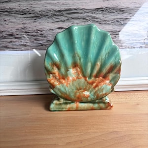 May include: A ceramic shell-shaped decorative object featuring a glossy finish with a blend of turquoise and burnt orange glaze. The piece has a scalloped edge and a flat base, showcasing a radial ribbed texture that mimics the natural form of a seashell.