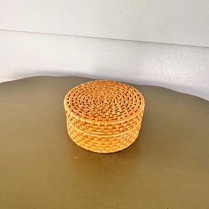 May include: A round, lidded storage box crafted from woven light brown rattan. The container features a tight, uniform basket-weave pattern across the flat lid and the cylindrical sides, showcasing a natural tan colour and a smooth, finished texture.