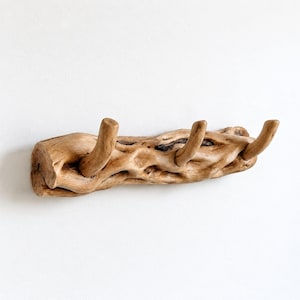 May include: A wall-mounted coat rack crafted from a single piece of light brown driftwood. The natural wood features a textured, gnarled surface with three integrated upward-curving hooks extending from the main horizontal branch structure.
