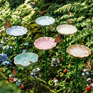 Può includere: Una collezione di sei vaschette per uccelli in ceramica su sottili steli metallici verdi, con bordi smerlati in tonalità pastello blu, rosa e beige. Due piccole statuette di rane grigie sono posizionate tra gli steli, con farfalle.