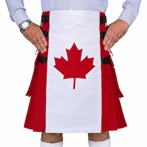 Op de afbeelding: Een rood-witte kilt met een opvallend rood esdoornbladontwerp gecentreerd op een wit voorpaneel. Het kledingstuk is voorzien van zwarte verstelbare zijbanden met gespen en geplooide rode stof aan de zijkanten, gedragen met een lichtblauw overhemd.
