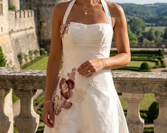 Könnte beinhalten: Ein cremeweißes Neckholder-Kleid mit geradem Ausschnitt und ausgestelltem Rock. Das Oberteil und der Rock sind mit gestickten Blumenapplikationen in Burgunderrot und Beige verziert, die sich von der rechten Schulter bis zur Hüfte erstrecken.