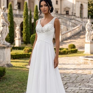 May include: A floor-length white sleeveless gown featuring a V-neckline with intricate silver beadwork and sequin embellishments on the bodice. The waist is defined by a wrapped pleated fabric detail, transitioning into a flowing A-line chiffon skirt.