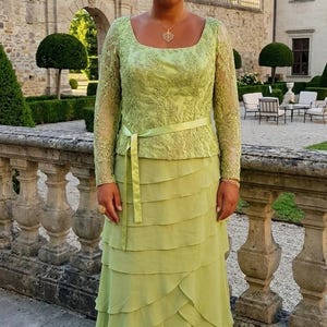 Könnte beinhalten: Ein limettengrünes, bodenlanges Kleid mit Rundhalsausschnitt, langen Spitzenärmeln mit aufwendiger Blumenstickerei und einem passenden Satinband als Gürtel in der Taille. Der Rock besteht aus mehreren horizontalen Stofflagen.