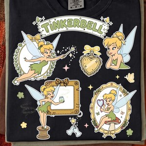 以下が含まれることがあります： 黒いTシャツに、杖を持ったり、鏡を見たり、糸巻きの上に立ったりしているティンカーベルのグラフィックプリントが施されています。デザインには、ハート型のロケット、額縁、そしてTinkerbellという文字が含まれています。