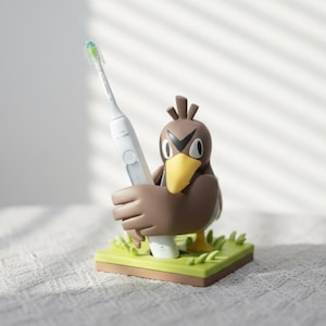 May include: A brown and yellow bird-shaped toothbrush holder standing on a square green base with moulded grass details. The holder features a cartoon-style bird with large eyes and a yellow beak, securely holding a white electric toothbrush.